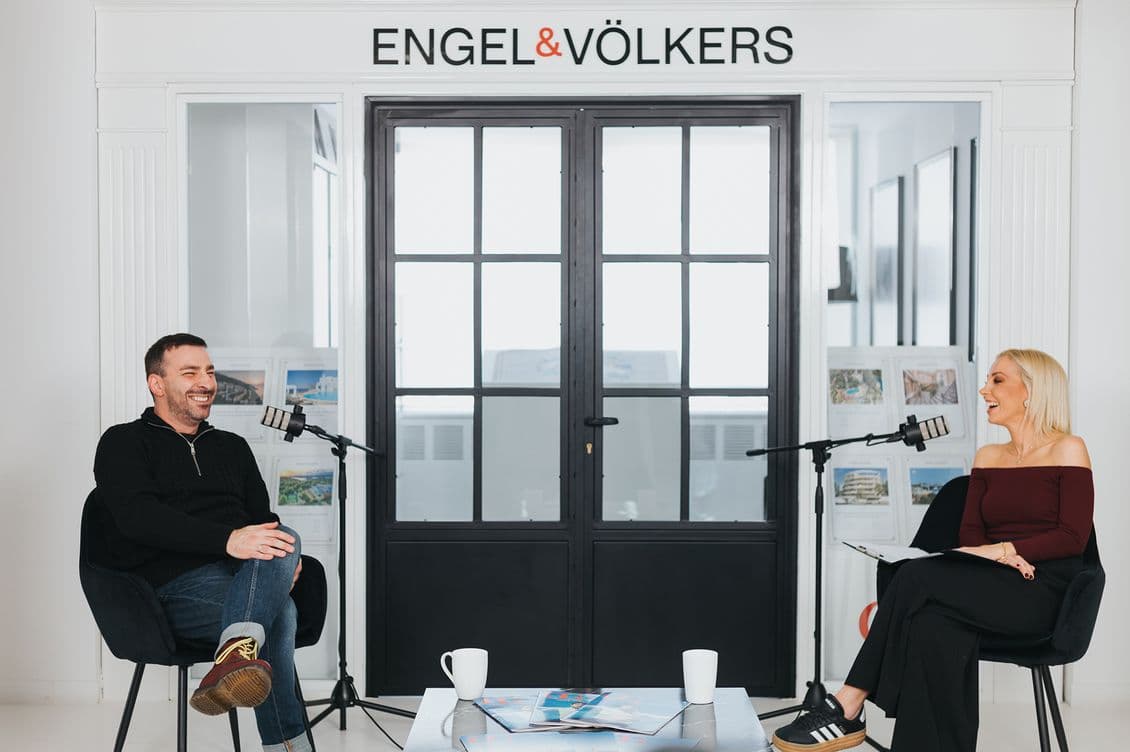 Two people seated with microphones, smiling in a modern office setting, with "Engel & Völkers" sign above. Coffee mugs on the table.