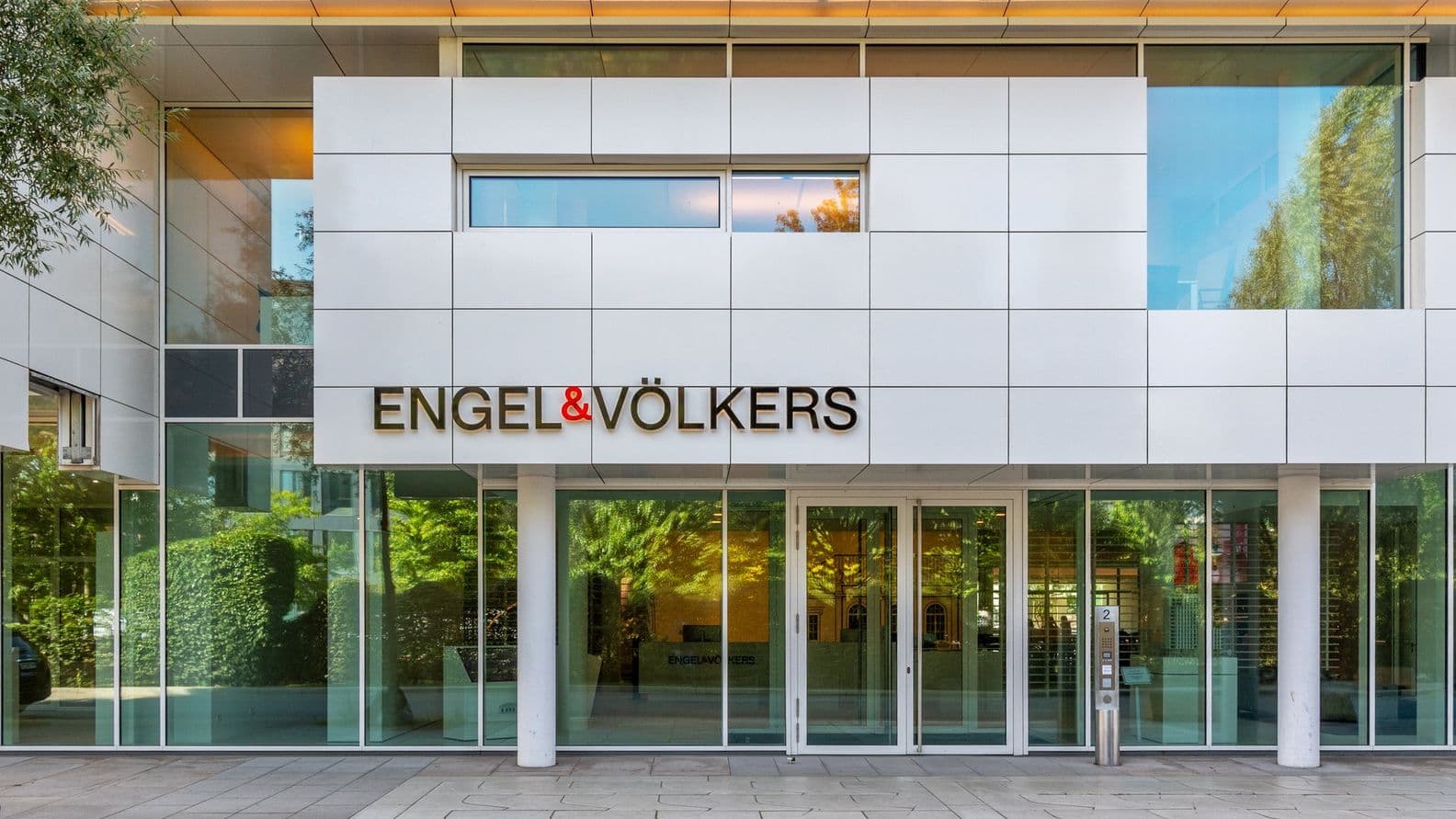 Moderne Gebäudefassade mit großen Fenstern und einem Schild mit der Aufschrift „ENGEL & VÖLKERS“ über dem Eingang.