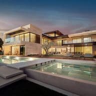 Modern luxury house with large glass windows, spacious balconies, and a pool, set against a dusk sky. Elegant landscaping surrounds the property.