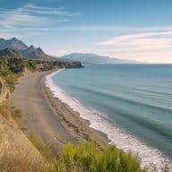 Curved sandy beach along the Mediterranean coast, with rugged cliffs, native vegetation, and a mountain range silhouetted in the distance.