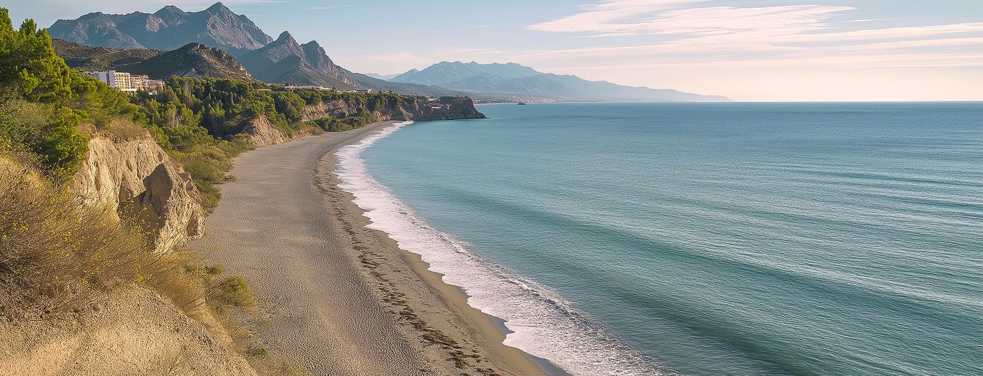 Platja de sorra corbada al llarg de la costa mediterrània, amb penya-segats, vegetació autòctona i una serralada muntanyosa perfilada a la distància.