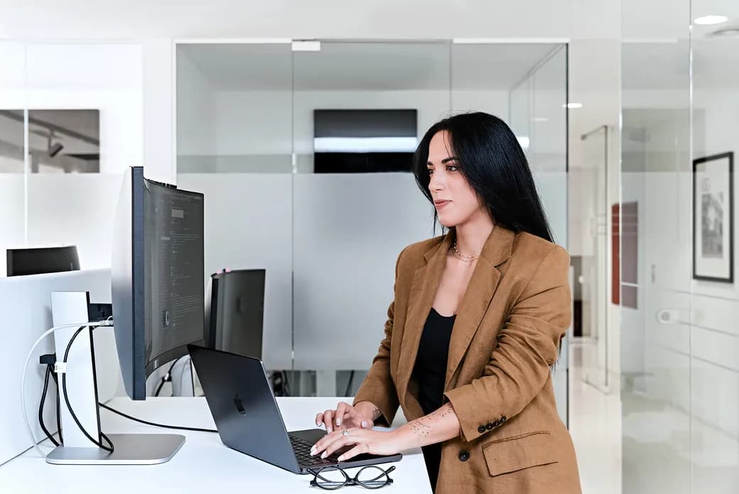 A professional woman with long dark hair in a tan blazer sits at a desk with a laptop and monitor in a modern glass-walled office.