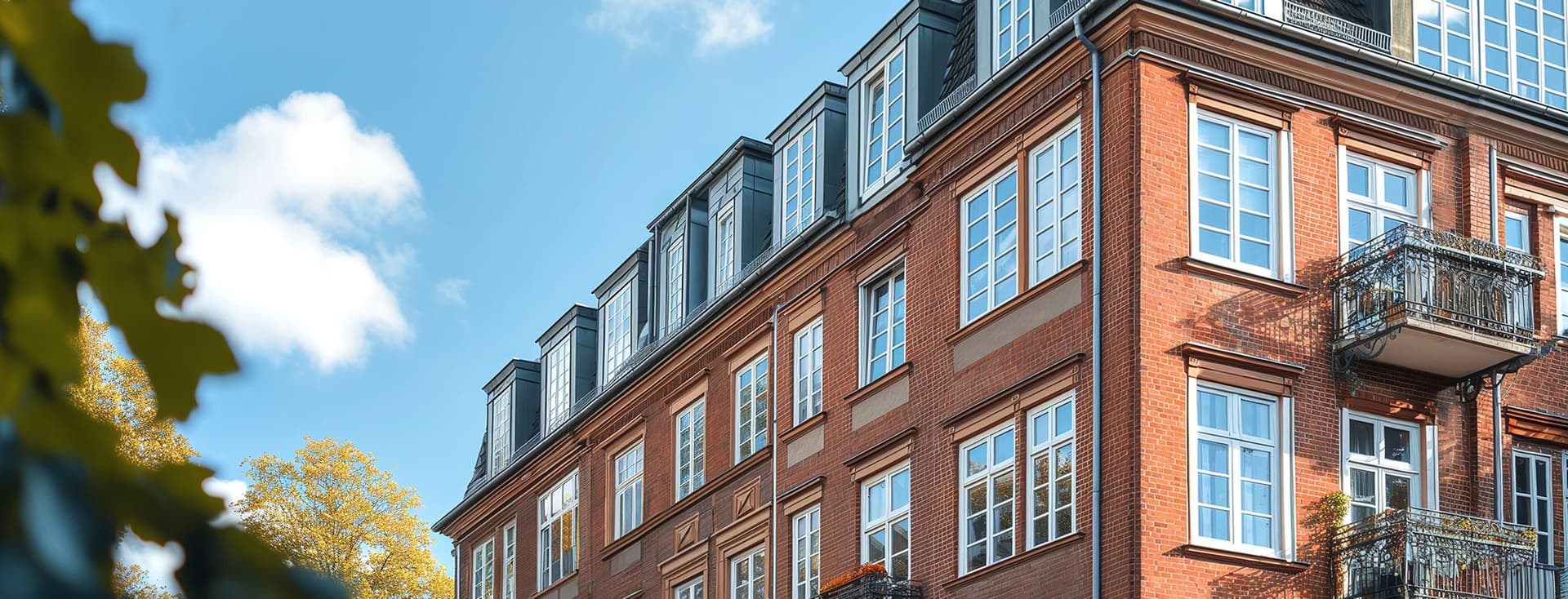 Außenansicht eines traditionellen Apartmentgebäudes aus rotem Backstein mit verzierten Balkonen, großen weiß gerahmten Fenstern und einem grauen Mansarddach mit Dachgauben. Herbstbäume umrahmen das Gebäude vor einem blauen Himmel mit weißen Wolken.