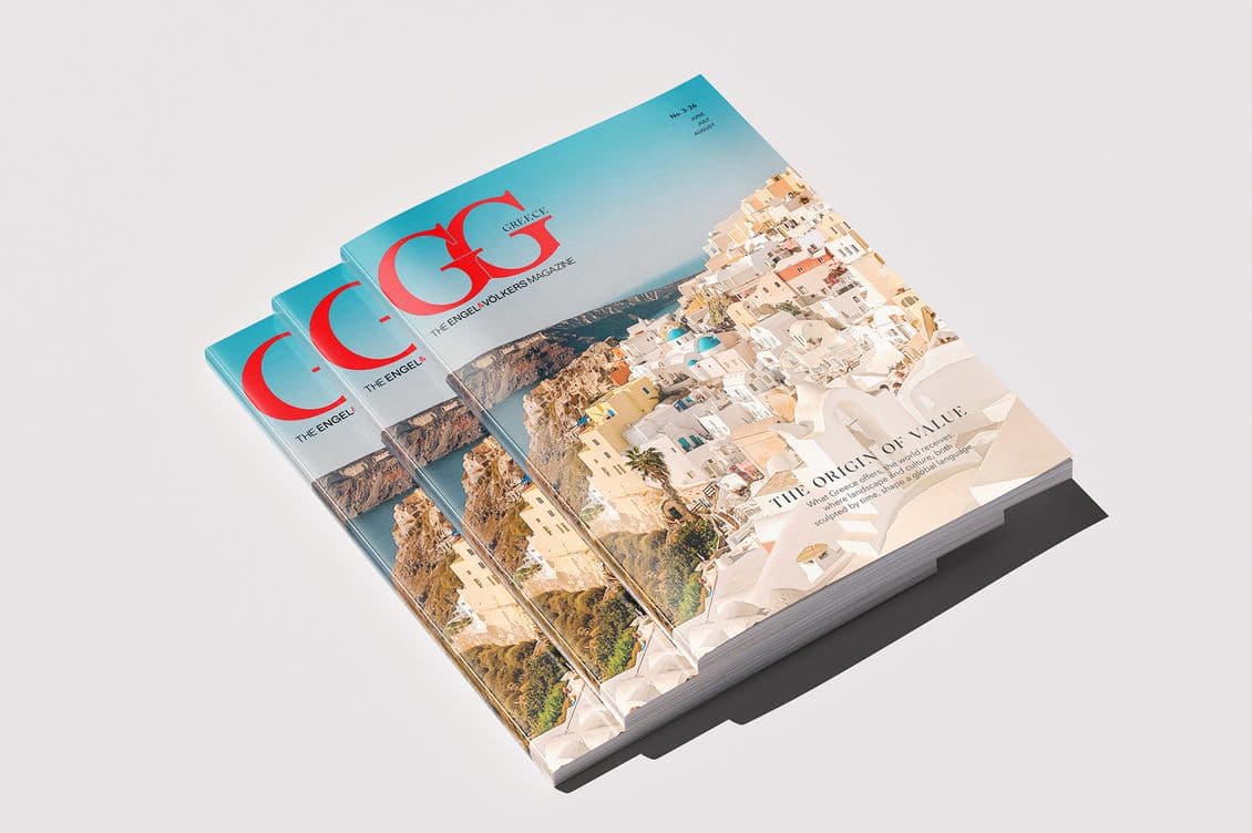 Stack of glossy Engel & Völkers Magazine issues with blue gradient covers, red logo, and a Santorini-style cliffside town photograph.