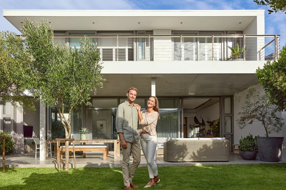Glückliches Paar im Garten ihrer finanzierten Immobilie – einem modernen, zweistöckigen Haus mit Balkon.