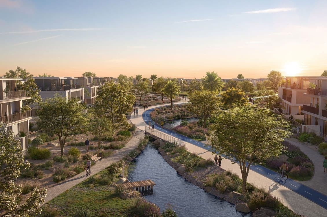 A scenic residential area at sunset features modern buildings, a winding path, and a tranquil stream lined with trees and plants.