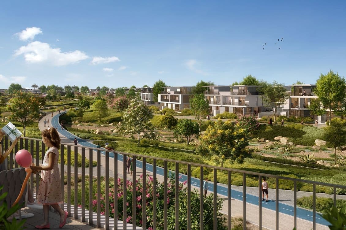 Woman and child with a balloon stand on a balcony overlooking a lush park with a winding path, trees, and modern buildings under a clear sky.