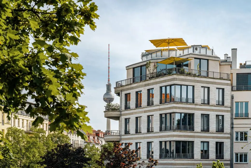 Modern multi-story building with yellow umbrellas on the rooftop, surrounded by trees, with a tower in the background under a clear sky.