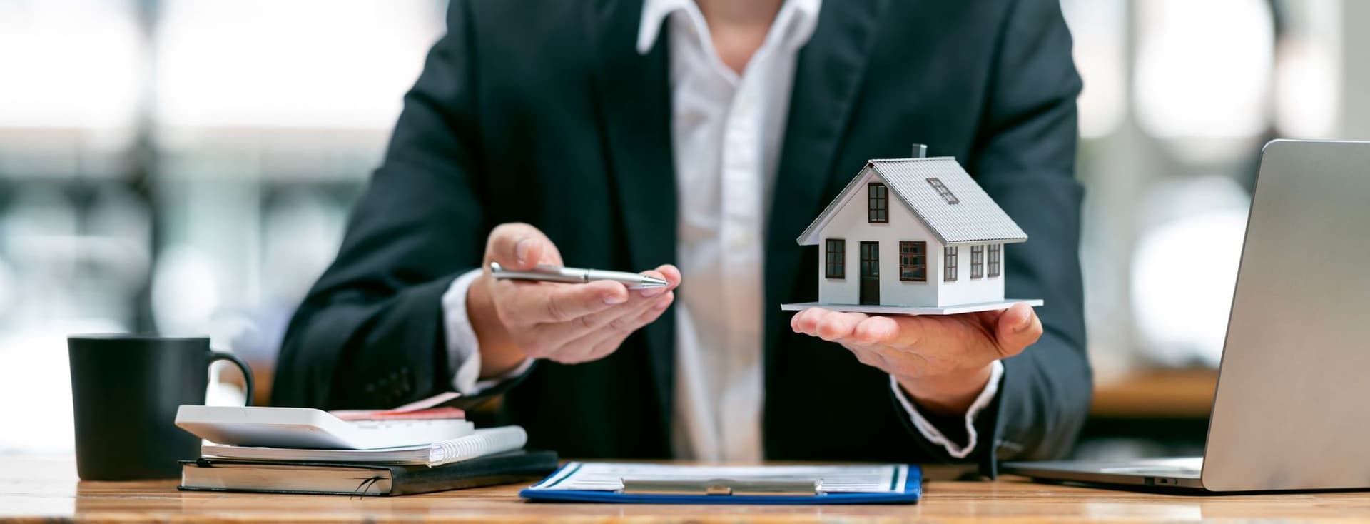 Businessman in a suit holding a model of a family house at his office desk, symbolizing value-based real estate investment and capital preservation.