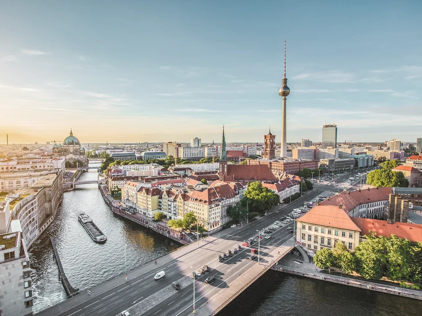 Panoramic view of Berlin at sunset overlooking the Spree River, with the Berlin Cathedral on the left, the Red City Hall, and the TV Tower in the center, surrounded by historic and modern buildings.