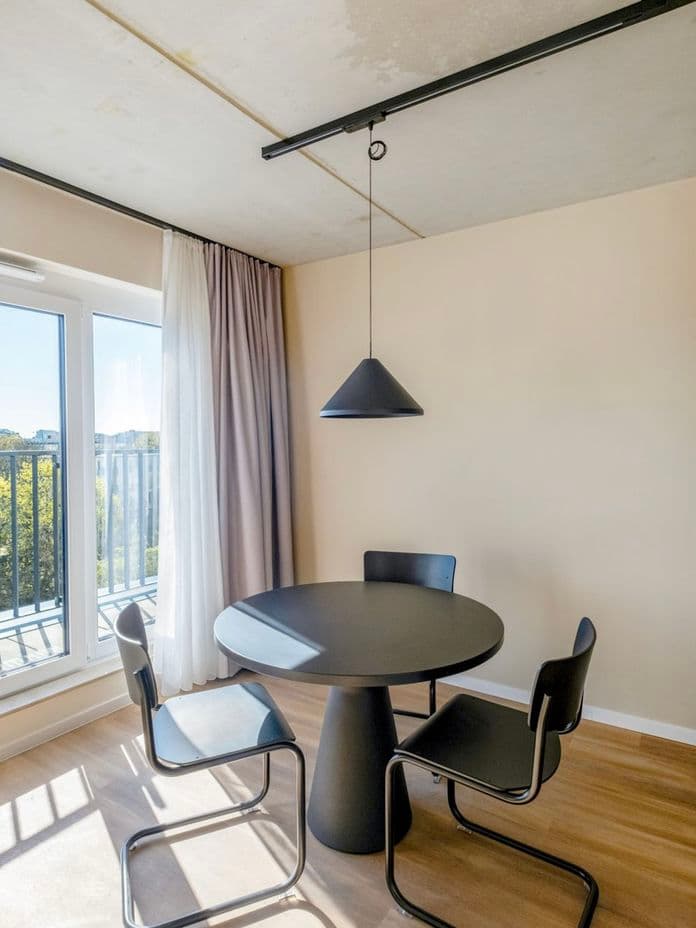 Modern dining area with a black round table, two black chairs, and pendant light. Floor-to-ceiling window with curtains and outside view.