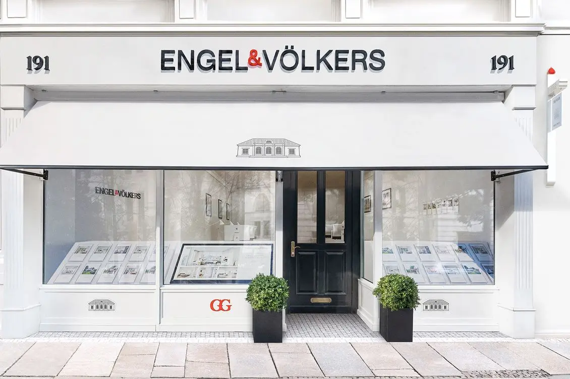 Front view of an Engel & Völkers real estate office with street number 191, featuring a minimalist white facade, central black entrance door flanked by two display windows showing property listings, black planters with boxwood spheres, and the company logo prominently displayed above with a classical villa illustration on the awning.
