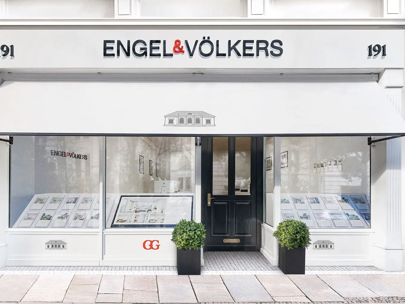 Front view of an Engel & Völkers real estate office with street number 191, featuring a minimalist white facade, central black entrance door flanked by two display windows showing property listings, black planters with boxwood spheres, and the company logo prominently displayed above with a classical villa illustration on the awning.