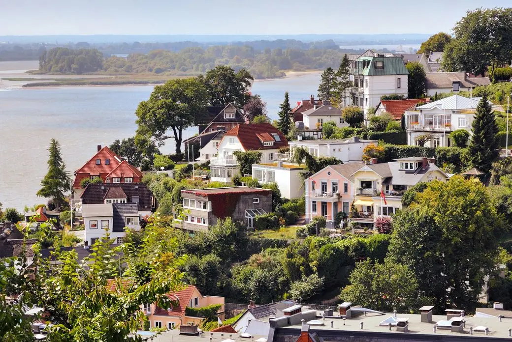 Scenic view of hillside houses with colorful roofs surrounded by greenery, overlooking a river and distant forested area.