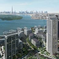 Aerial view of a modern cityscape with waterfront buildings, a river, and a distant skyline featuring a tall tower under a clear blue sky.
