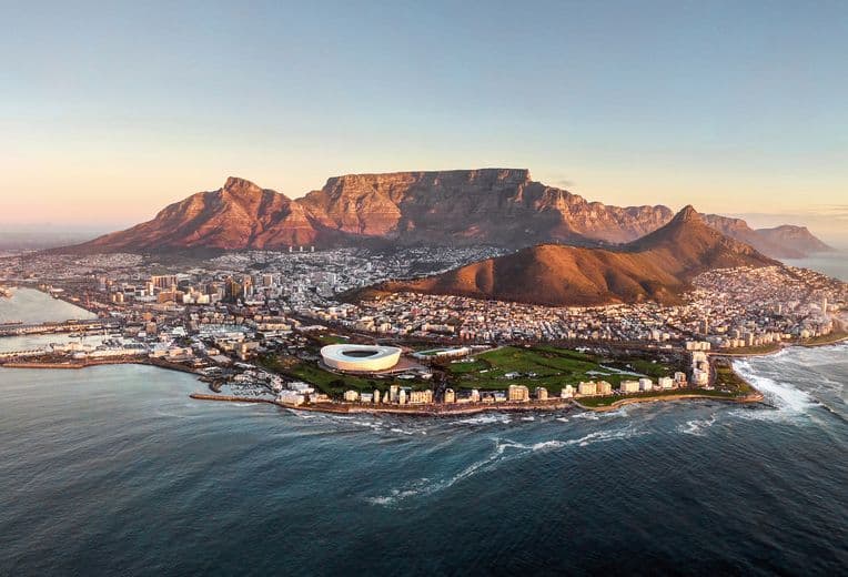 Luftaufnahme von Kapstadt, Südafrika, mit dem ikonischen Tafelberg im Hintergrund bei Sonnenaufgang. Die Stadt breitet sich entlang der Küste aus, mit dem Kapstadt-Stadion und der städtischen Bebauung zwischen dem Berg und dem Atlantischen Ozean, wo sich die Wellen an der Küste brechen.