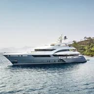 Luxury superyacht anchored near a Mediterranean coastline, featuring a sleek grey and white exterior design with multiple decks, satellite communications equipment, and panoramic windows. The vessel is photographed against a backdrop of green hillsides and calm blue waters.