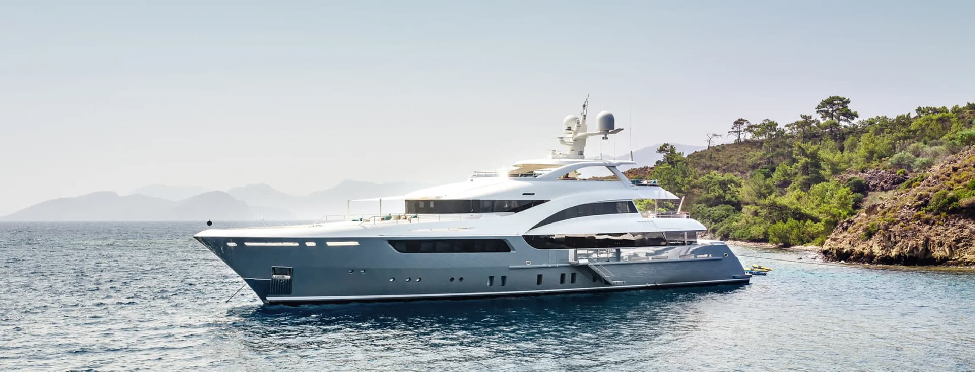 Luxury superyacht anchored near a Mediterranean coastline, featuring a sleek grey and white exterior design with multiple decks, satellite communications equipment, and panoramic windows. The vessel is photographed against a backdrop of green hillsides and calm blue waters.
