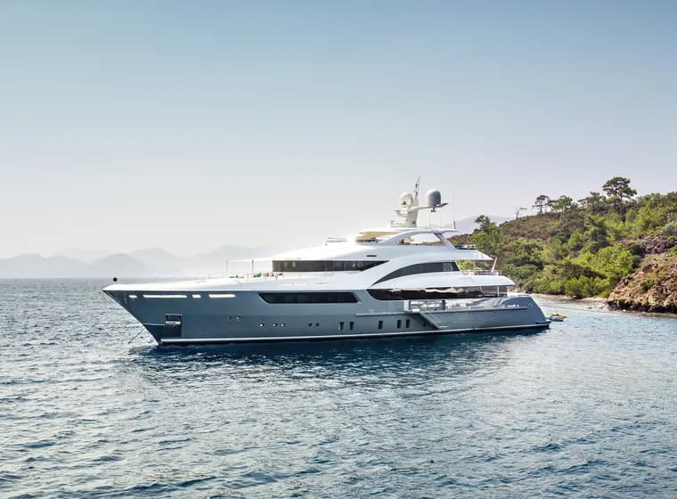 Luxury superyacht anchored near a Mediterranean coastline, featuring a sleek grey and white exterior design with multiple decks, satellite communications equipment, and panoramic windows. The vessel is photographed against a backdrop of green hillsides and calm blue waters.