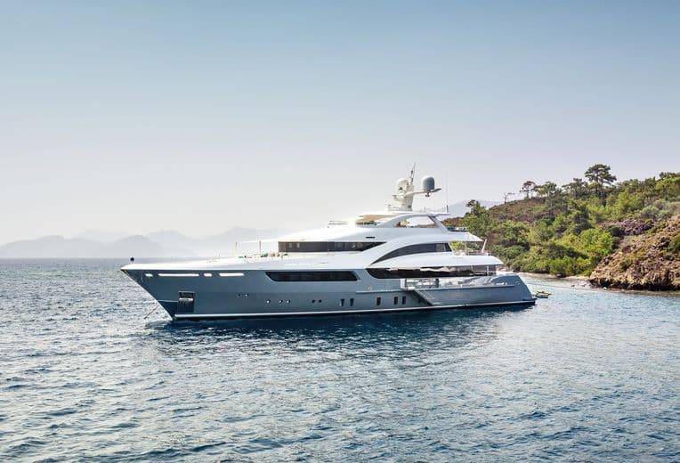 Luxuriöse Superyacht, die in der Nähe der Mittelmeerküste vor Anker liegt und ein elegantes, grau-weißes Außendesign mit mehreren Decks, Satellitenkommunikationsausrüstung und Panoramafenstern aufweist. Das Schiff wurde vor einer Kulisse aus grünen Hängen und ruhigem, blauem Wasser fotografiert.