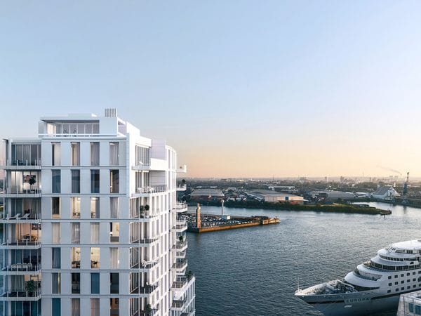 Aerial view of a modern white apartment building with multiple balconies overlooking a harbor in Hamburg. A cruise ship is docked nearby, and the cityscape extends into the distance under a clear sky at dusk, highlighting the premium waterfront real estate location.