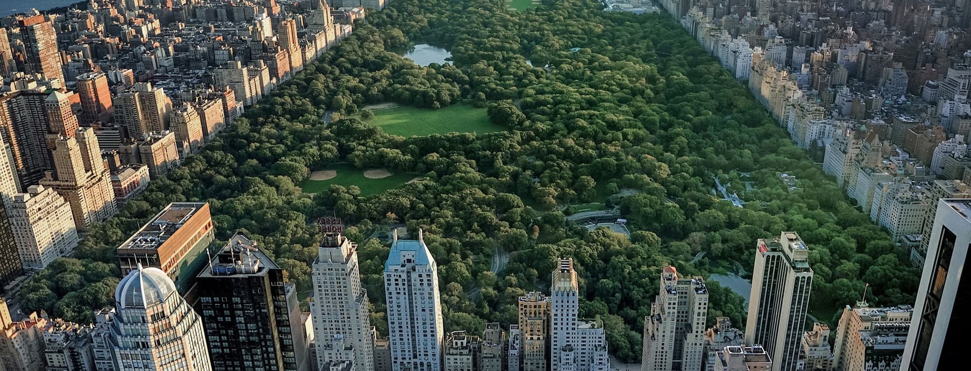 Aerial view of New York City's Central Park, showing the vast rectangular green space surrounded by Manhattan's towering skyscrapers. The park's lush trees and lawns contrast dramatically with the dense urban landscape, with high-rise buildings lining both sides and water bodies visible in the distance.