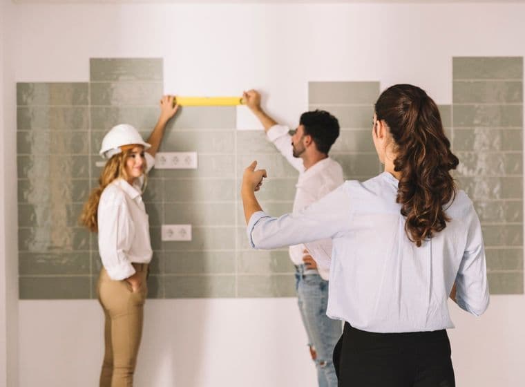 Three people measure tile on a wall. One woman wears a hard hat. A woman points at the wall. A man holds a yellow level.