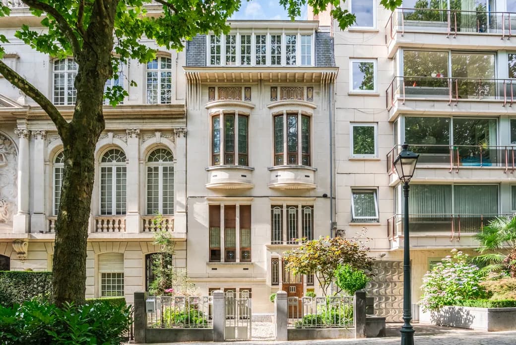 Historic beige stone building with ornate bay windows and balconies beside a modern glass building; a large tree and street lamp frame the scene. Image générée par l'IA