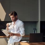 Man in a white shirt stands in a kitchen, drinking coffee and reading a magazine, with a laptop and sunlight streaming in.
