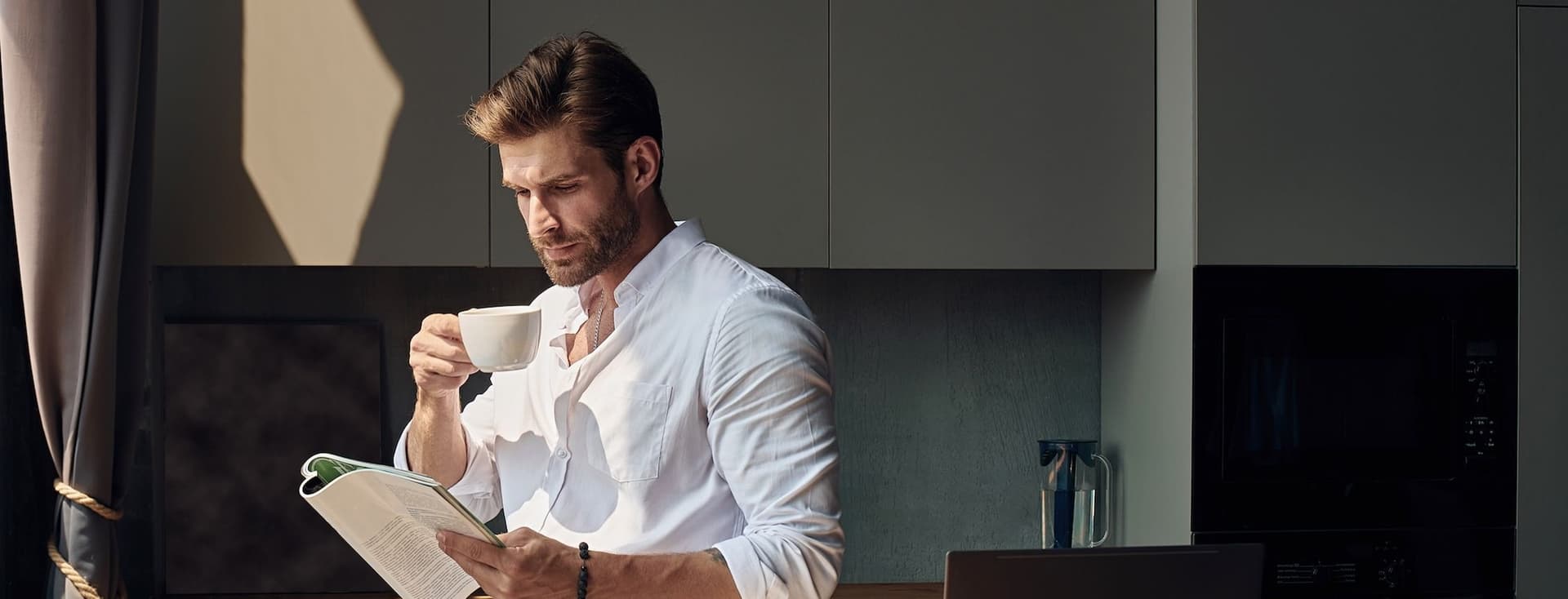 Man in a white shirt stands in a kitchen, drinking coffee and reading a magazine, with a laptop and sunlight streaming in.