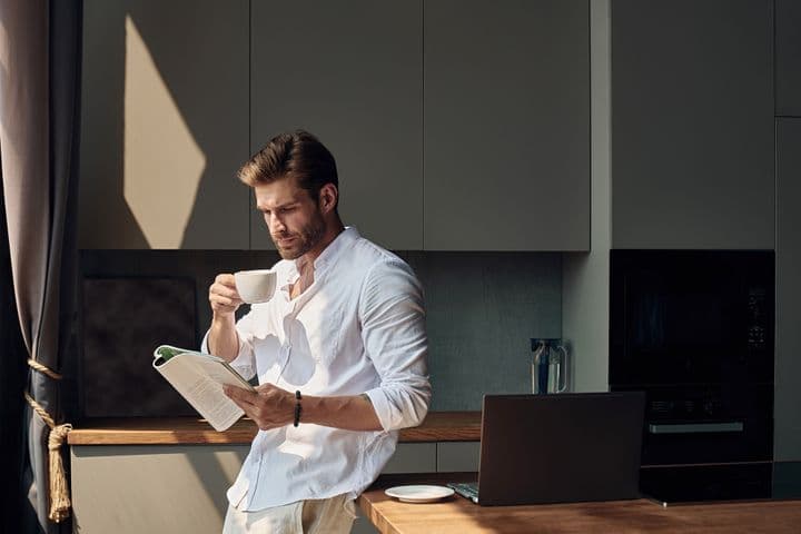 Man in a white shirt stands in a kitchen, drinking coffee and reading a magazine, with a laptop and sunlight streaming in.