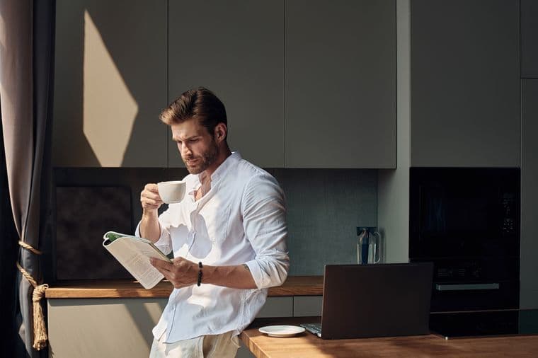 Man in a white shirt stands in a kitchen, drinking coffee and reading a magazine, with a laptop and sunlight streaming in.