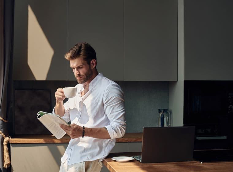 Man in a white shirt stands in a kitchen, drinking coffee and reading a magazine, with a laptop and sunlight streaming in.
