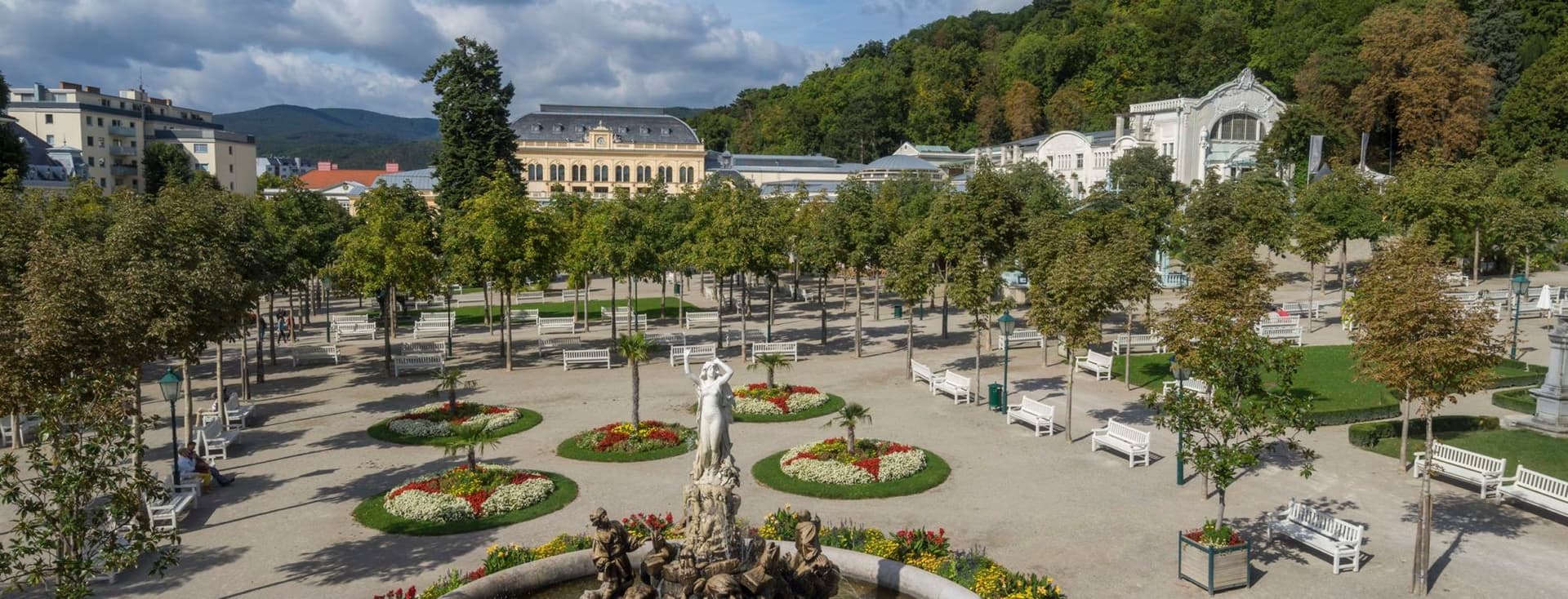 Weitläufiger Kurpark mit historischem Springbrunnen im Zentrum, umgeben von symmetrisch angelegten Blumenbeeten, weißen Parkbänken und Alleebäumen, im Hintergrund das Kurhaus