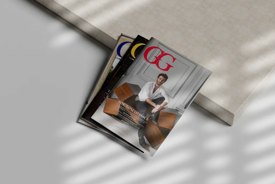 Multiple copies of GG Magazine stacked at an angle on a light surface, featuring a cover with a person in white attire in an elegant interior setting with wooden furniture