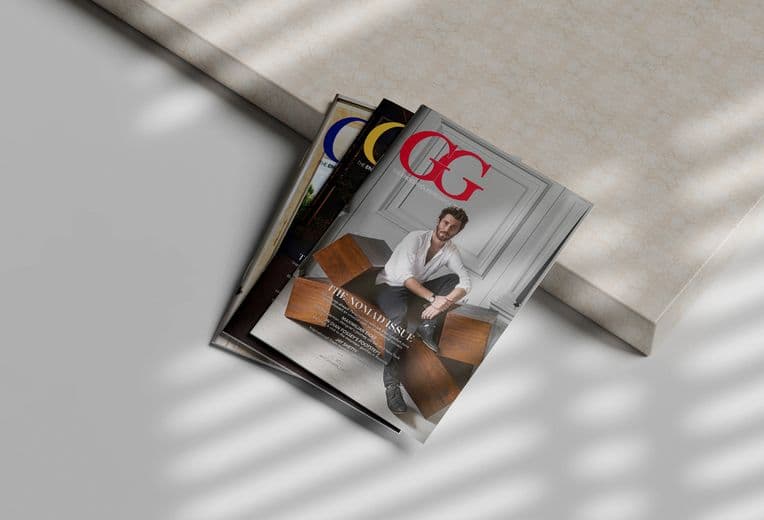 Multiple copies of GG Magazine stacked at an angle on a light surface, featuring a cover with a person in white attire in an elegant interior setting with wooden furniture