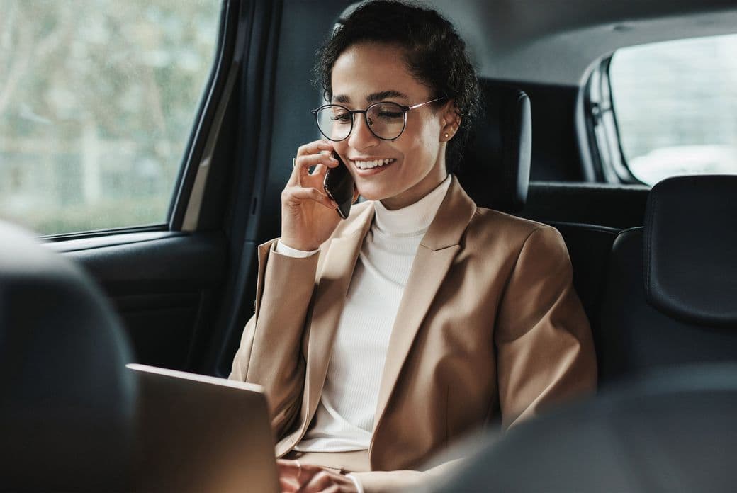 A professional woman wearing glasses, a beige blazer, and white turtleneck sits in the back seat of a car, smiling while talking on her phone and looking at a laptop. She appears to be conducting business while traveling.