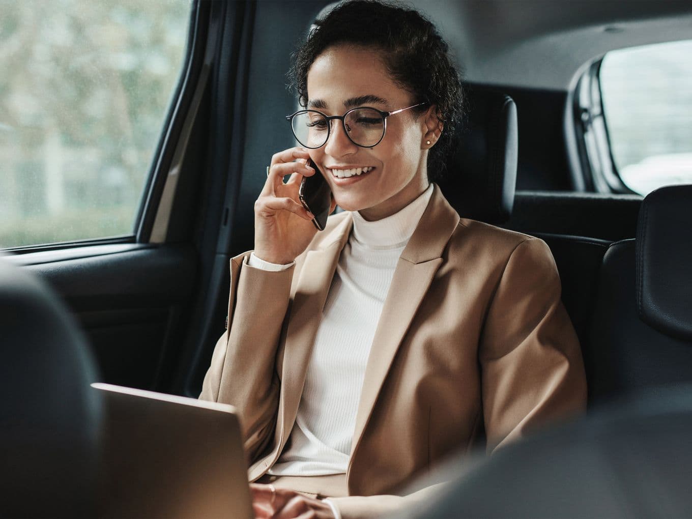 A smiling woman in a tan blazer and glasses talks on her phone while working on a laptop in the back of a car.