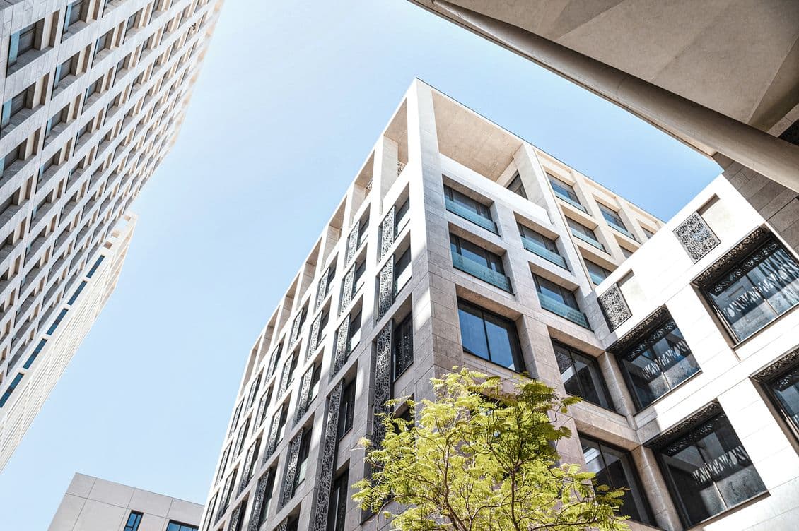 Prise de vue en contre-plongée de bâtiments modernes en pierre claire sur fond de ciel bleu, avec un arbre au premier plan.