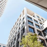 Upward view of a modern commercial building with geometric white facade, featuring repeating window patterns, glass elements, and a young tree in the foreground against a blue sky