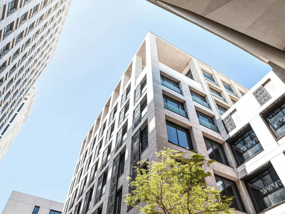 Low-angle shot of modern, light-stone buildings against a blue sky, with a tree in the foreground.