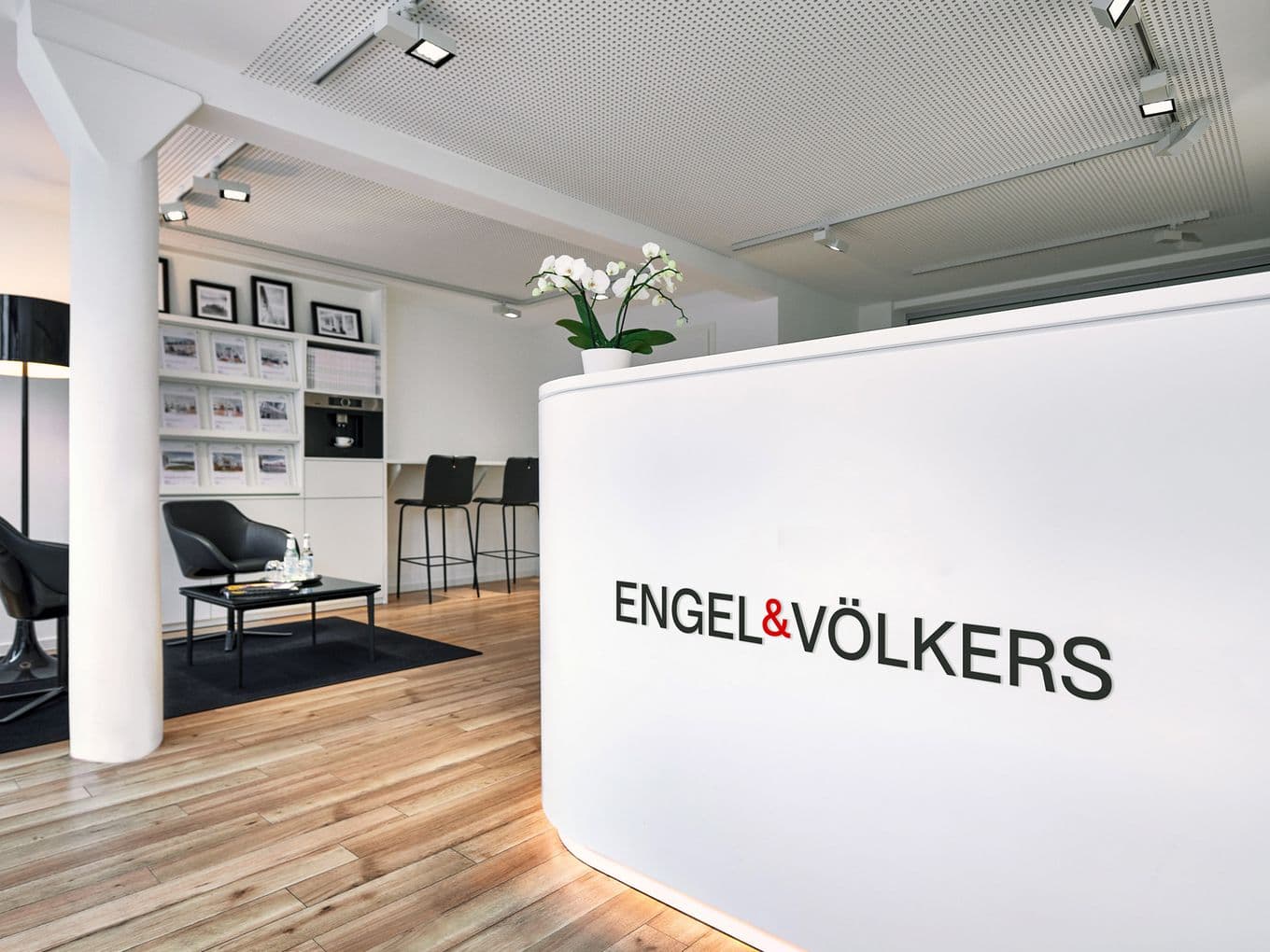 Engel & Völkers office reception area with a white desk, orchid, and seating. The logo is displayed on the desk.