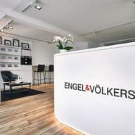 Innenansicht eines Engel & Völkers-Büros in Eimsbüttel mit einem geschwungenen weißen Empfangstresen mit beleuchtetem Logo, einer schwarzen Ledersitzecke mit Stehlampe, einer Immobilienanzeigewand und einer modernen industriellen Decke mit Strahlern. Der Raum kombiniert einen natürlichen Holzboden mit einem klaren schwarz-weißen Designschema.