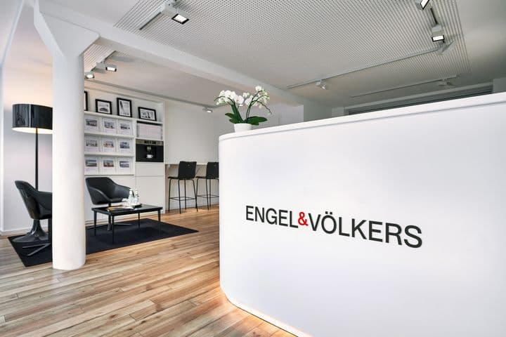 Innenansicht eines Engel & Völkers-Büros in Eimsbüttel mit einem geschwungenen weißen Empfangstresen mit beleuchtetem Logo, einer schwarzen Ledersitzecke mit Stehlampe, einer Immobilienanzeigewand und einer modernen industriellen Decke mit Strahlern. Der Raum kombiniert einen natürlichen Holzboden mit einem klaren schwarz-weißen Designschema.