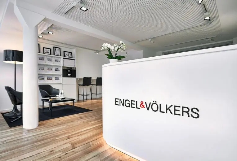 nterior view of an Engel & Völkers office in Eimsbuettel featuring a curved white reception counter with illuminated logo, black leather seating area with floor lamp, property display wall, and modern industrial ceiling with spotlights. The space combines natural wood flooring with a clean black and white design scheme.
