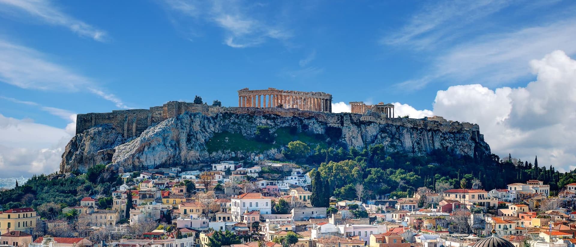 Die Athener Stadtlandschaft mit der Akropolis auf einem felsigen Hügel unter blauem Himmel mit vereinzelten Wolken. Gebäude mit roten Ziegeldächern füllen den Vordergrund.
