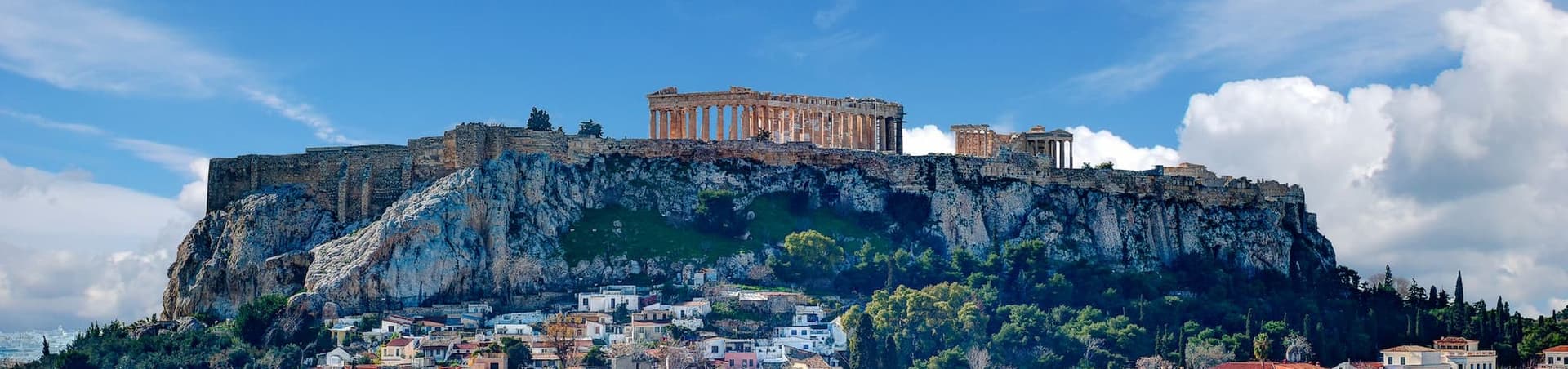 Athens cityscape with the Acropolis on a rocky hill under a blue sky with scattered clouds. Buildings with red tile roofs fill the foreground.