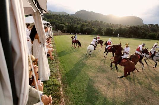 Polo players in white uniforms competing on horseback during a match on a lush green field, viewed from a spectator tent with mountains in the background at sunset