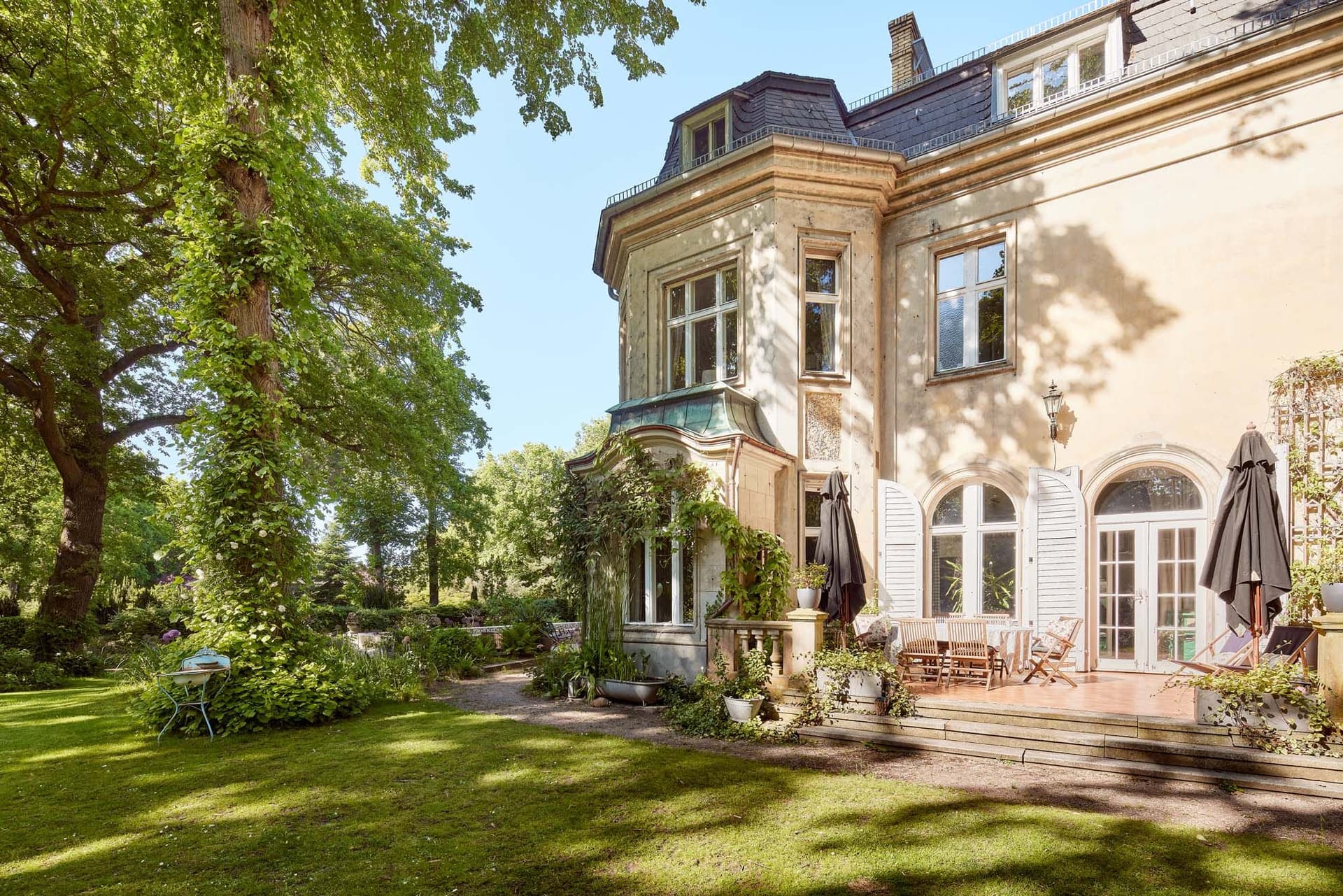 Historic mansion with large windows, surrounded by lush greenery and a spacious lawn, with a terrace equipped with chairs and parasols.