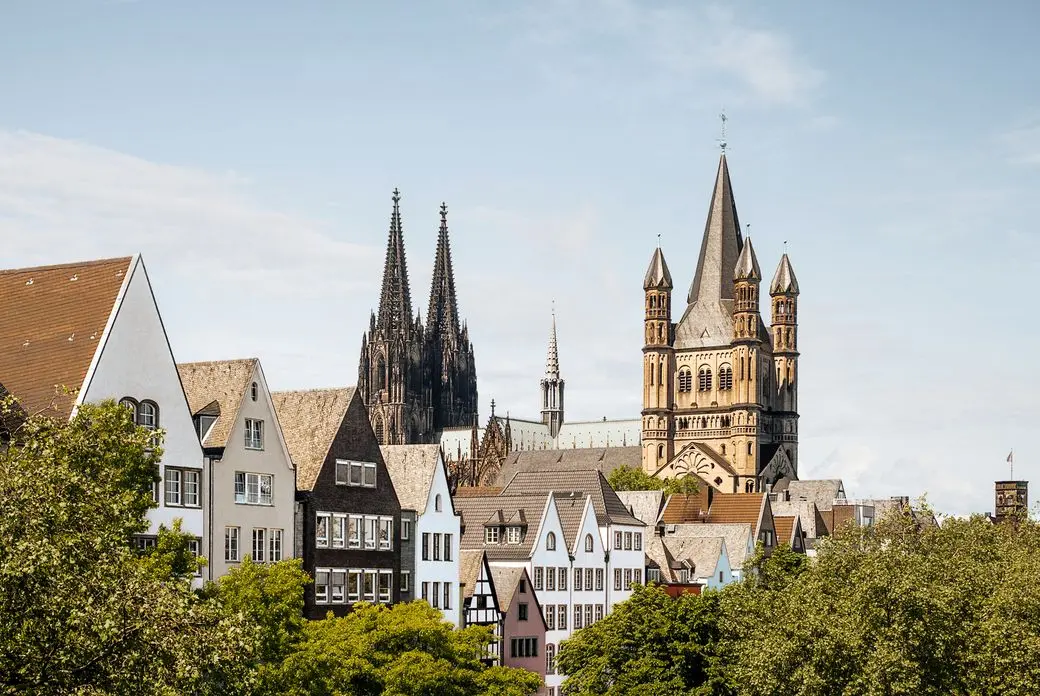 Skyline of Cologne featuring the Gothic Cologne Cathedral, Romanesque Great St. Martin Church, and traditional colorful buildings in the foreground.